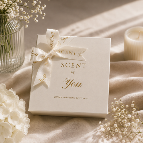 A white gift box tied with a cream satin ribbon printed with 'SCENT of You' in gold lettering, styled on draped cream fabric with white flowers and a candle in the background.